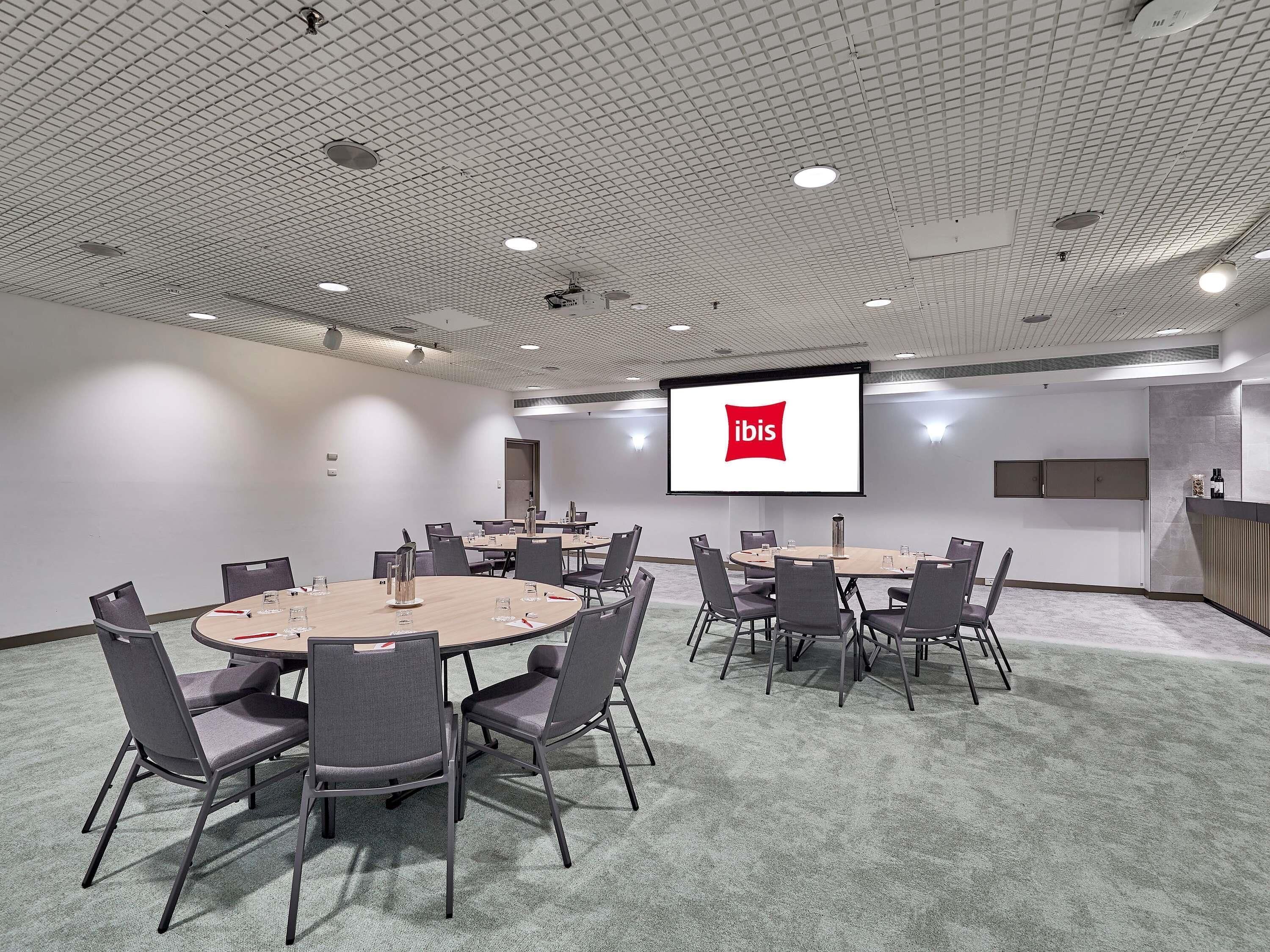 A conference room with a large screen displaying the ibis logo, multiple tables with chairs, and a kitchenette area.