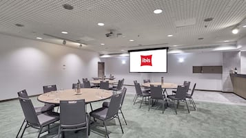 A conference room with a large screen displaying the ibis logo, multiple tables with chairs, and a kitchenette area.