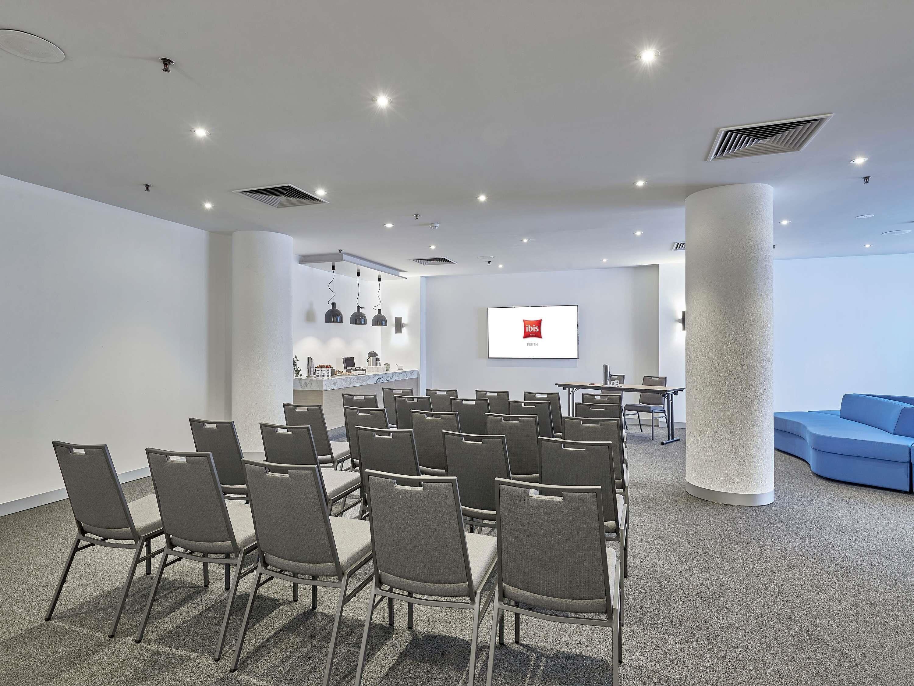 Ein Konferenzraum mit grauen Stühlen, einem weißen Tisch, einem Bildschirm mit dem ibis-Logo und einem blauen Sofa.