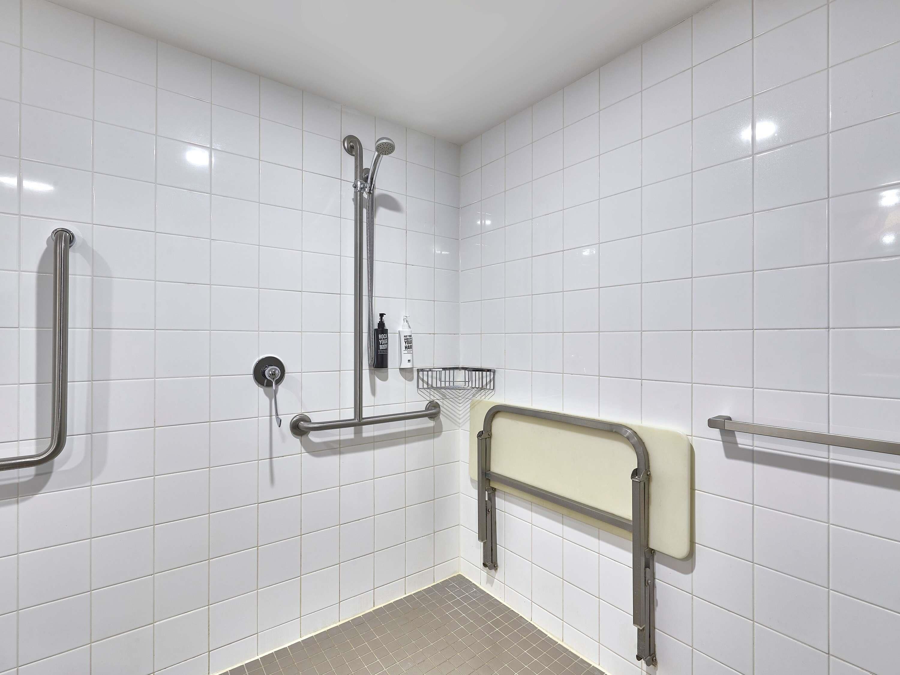 A bathroom with white tiled walls, a shower with a handheld showerhead, a grab bar, and a toilet with a safety bar.