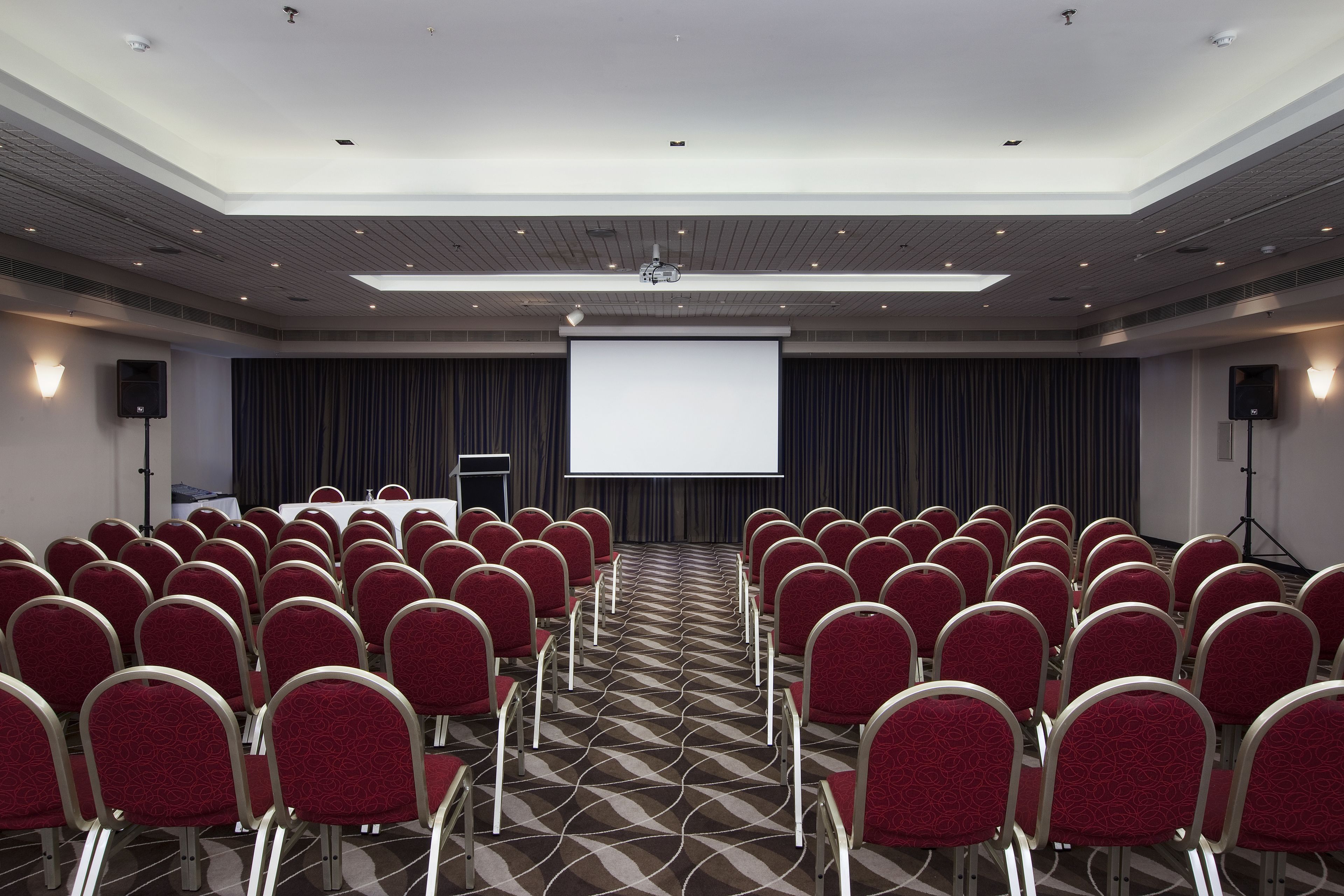 A conference room with rows of red chairs, a large screen, and a podium.