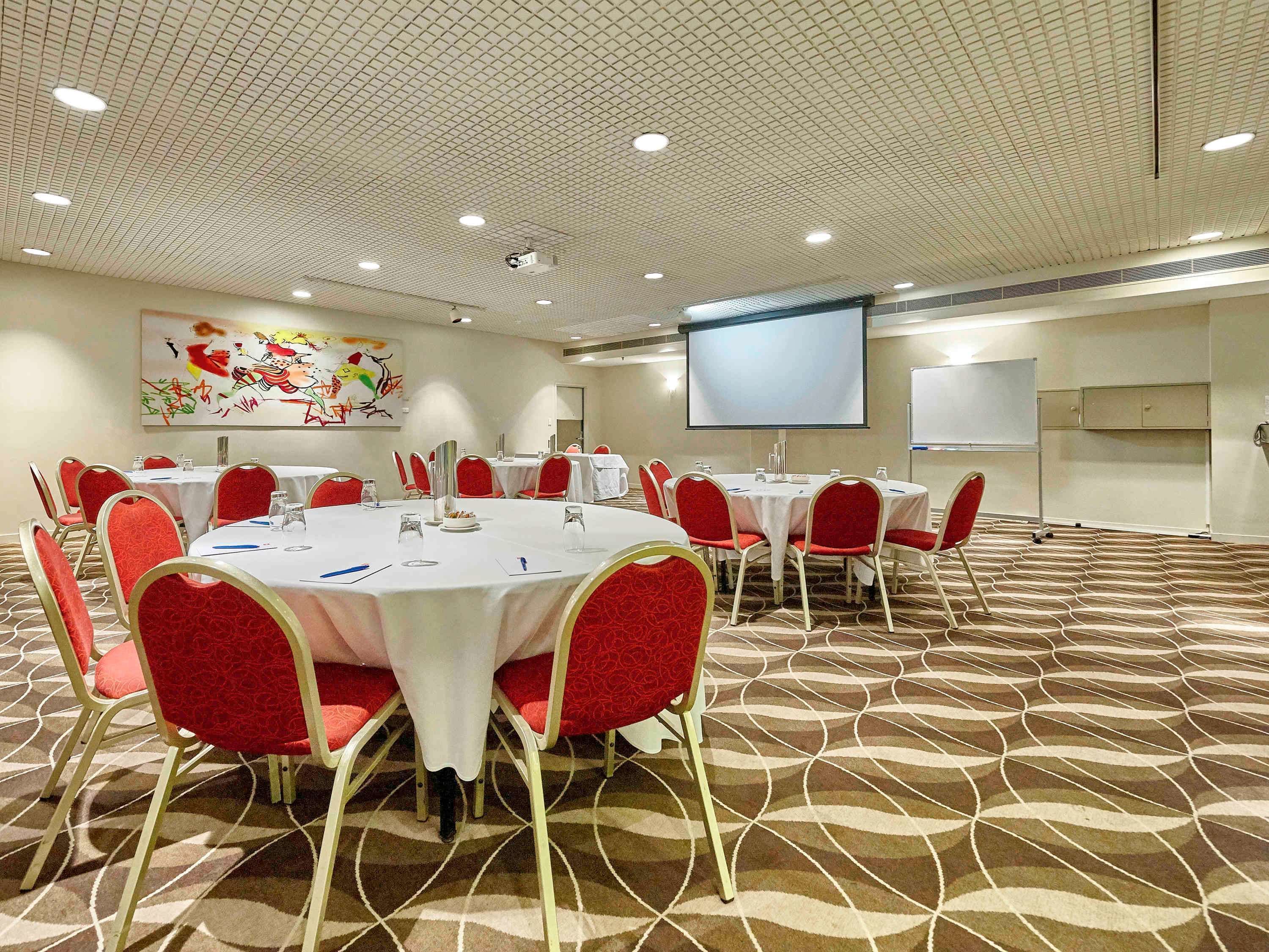 Uma sala de conferência com mesas redondas, cadeiras vermelhas, uma tela de projetor e um quadro branco.