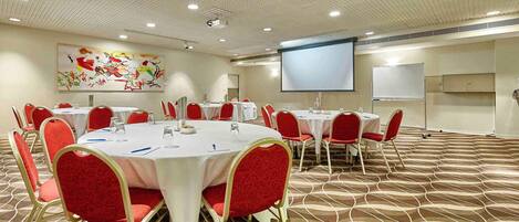 A conference room with round tables, red chairs, a projector screen, and a whiteboard.