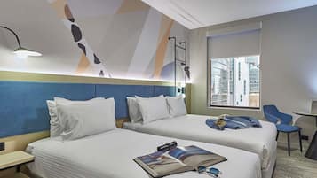 A hotel room with two beds, a desk, a chair, a window with a city view, and a modern abstract wall design.