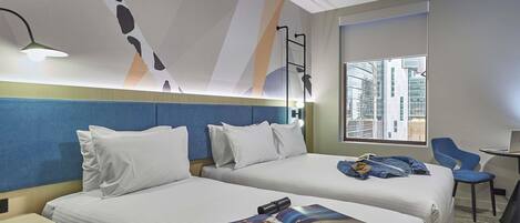 A hotel room with two beds, a desk, a chair, a window with a city view, and a modern abstract wall design.