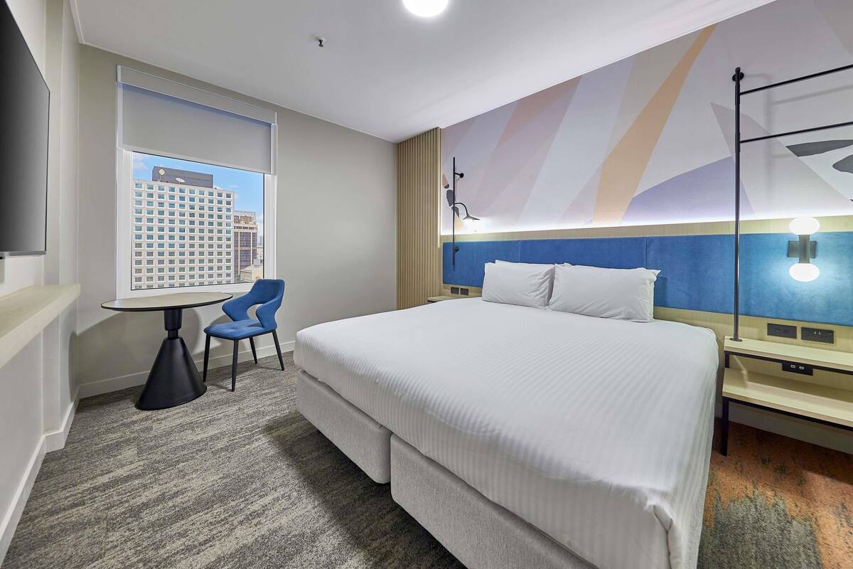 A hotel room with a bed, a desk, a chair, a TV, and a window with a city view.