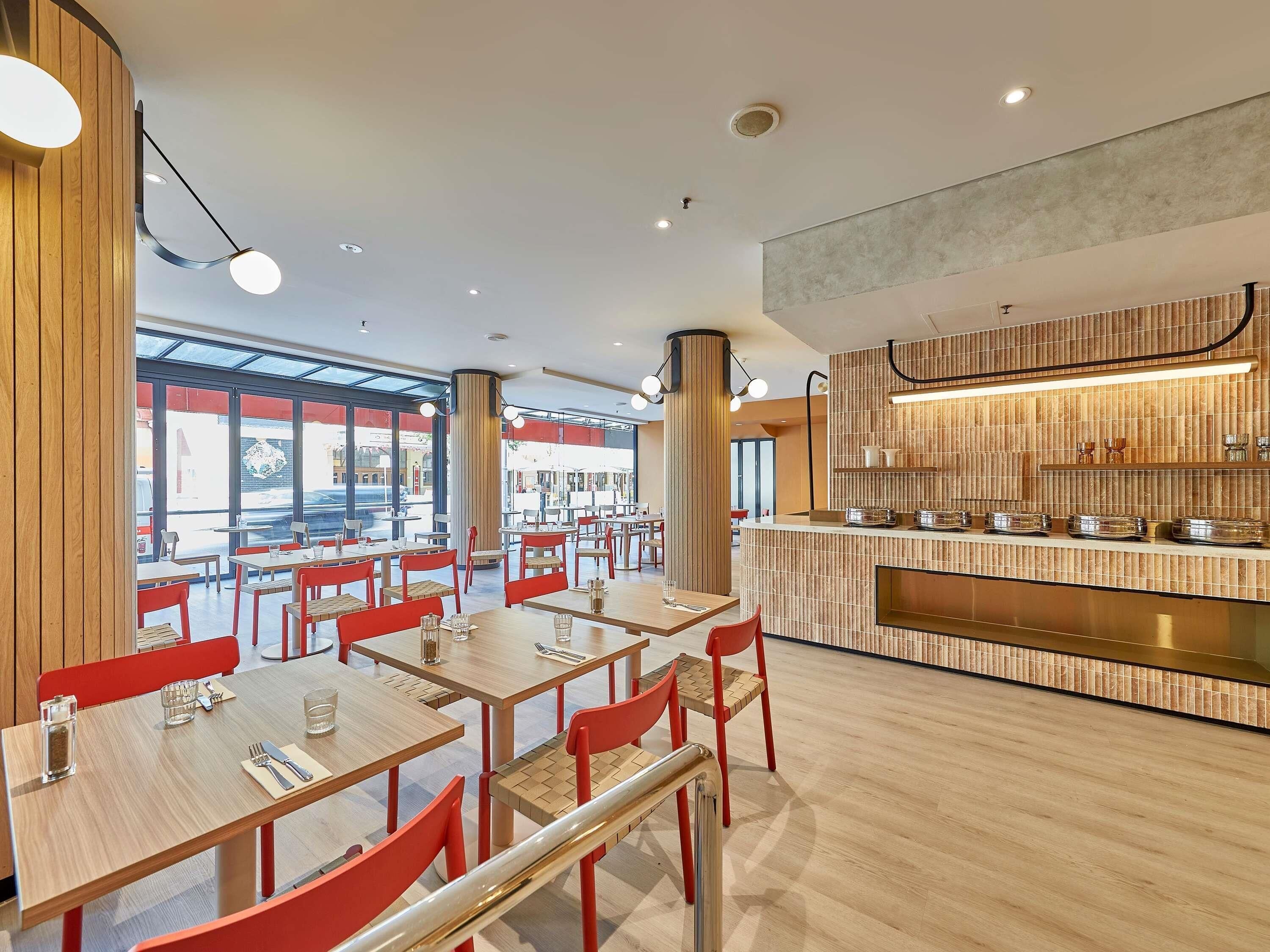 A modern restaurant interior with wooden tables, red chairs, and a central counter with a tiled backsplash.