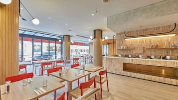 A modern restaurant interior with wooden tables, red chairs, and a central counter with a tiled backsplash.