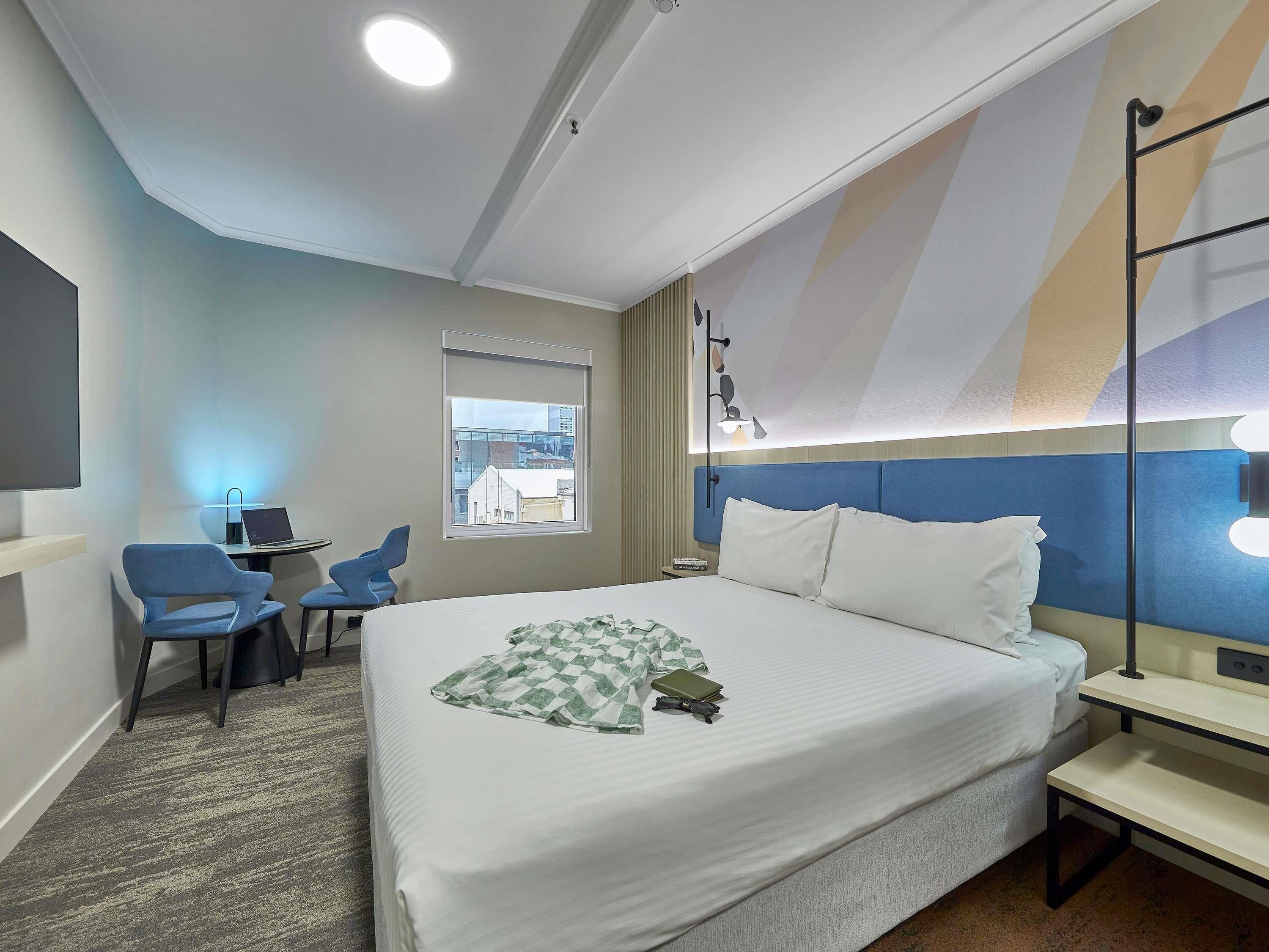 A hotel room with a bed, a desk, a chair, and a window with a view of buildings.