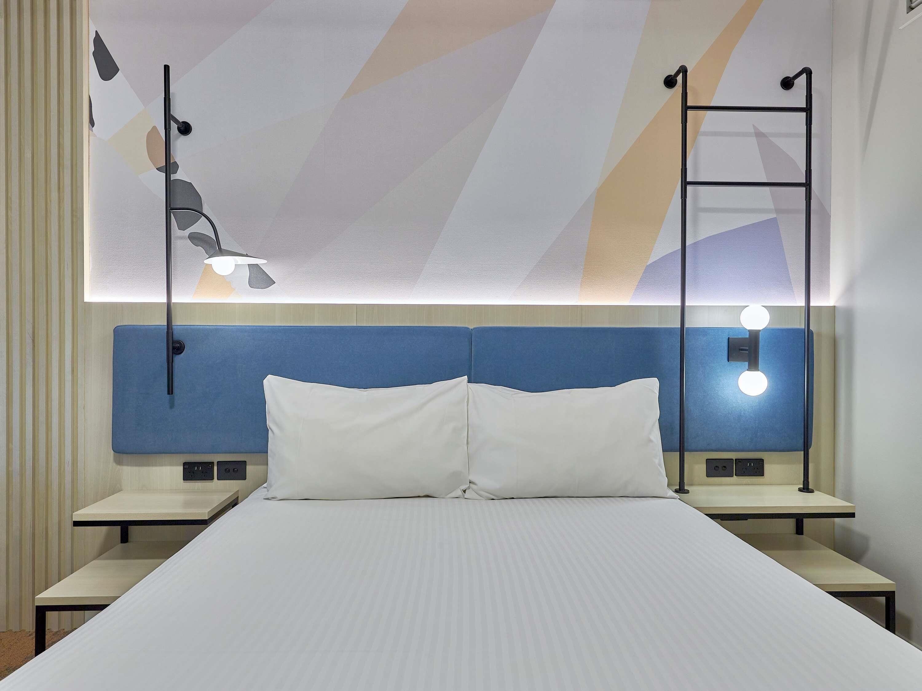 Een hotelkamer met een groot bed, twee nachtkastjes, een blauw hoofdeinde en een wandlamp.