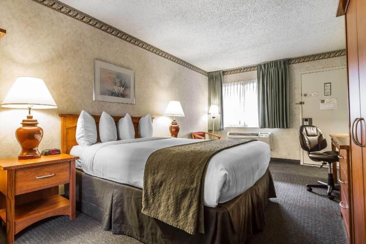Ein Hotelzimmer mit einem Bett, zwei Nachttischlampen, einem Schreibtisch mit Lampe, einem Sessel, einer Kommode, einer Klimaanlage und einem Fenster mit Vorhängen.