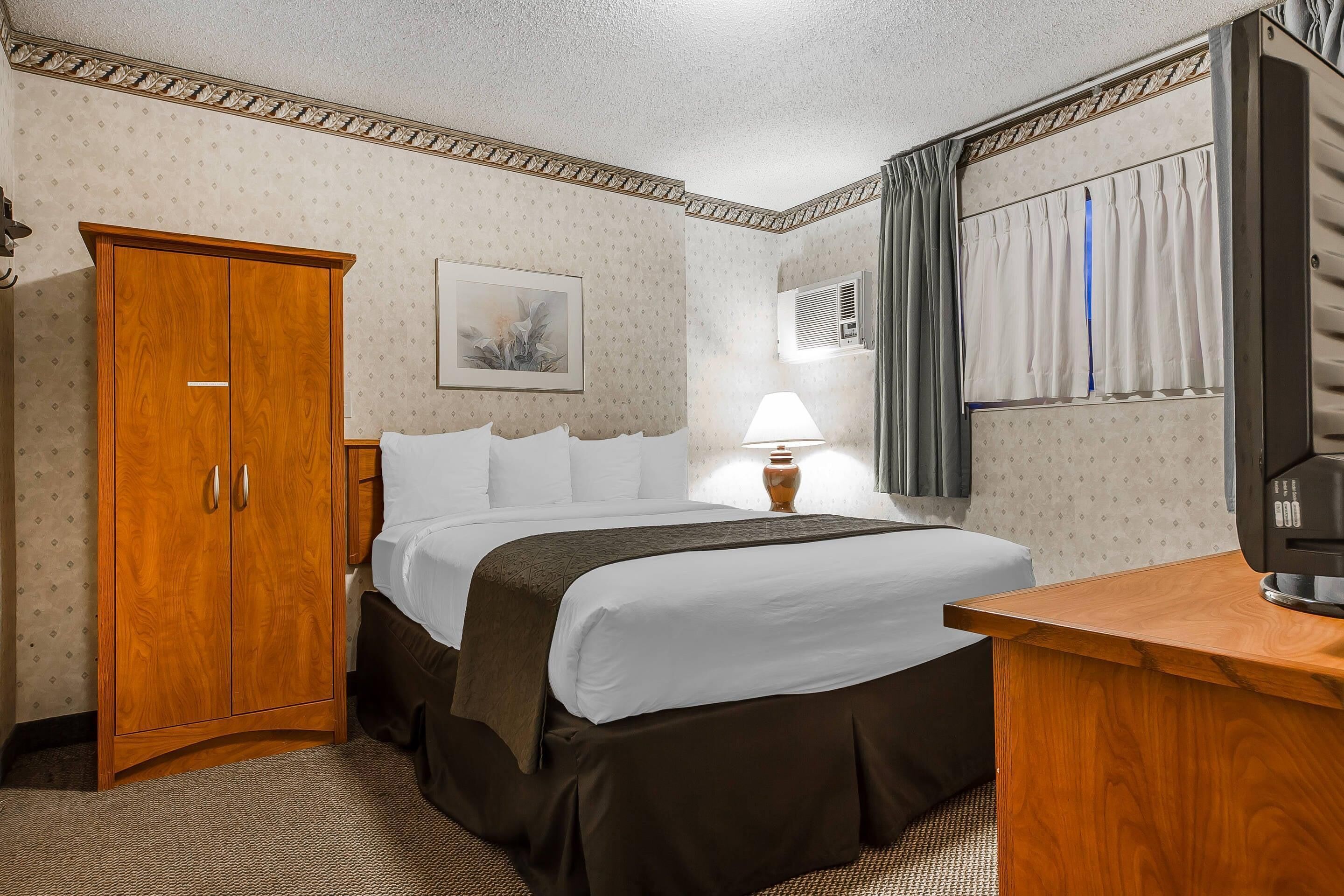 A hotel room with a bed, a wooden wardrobe, a desk, and a lamp.