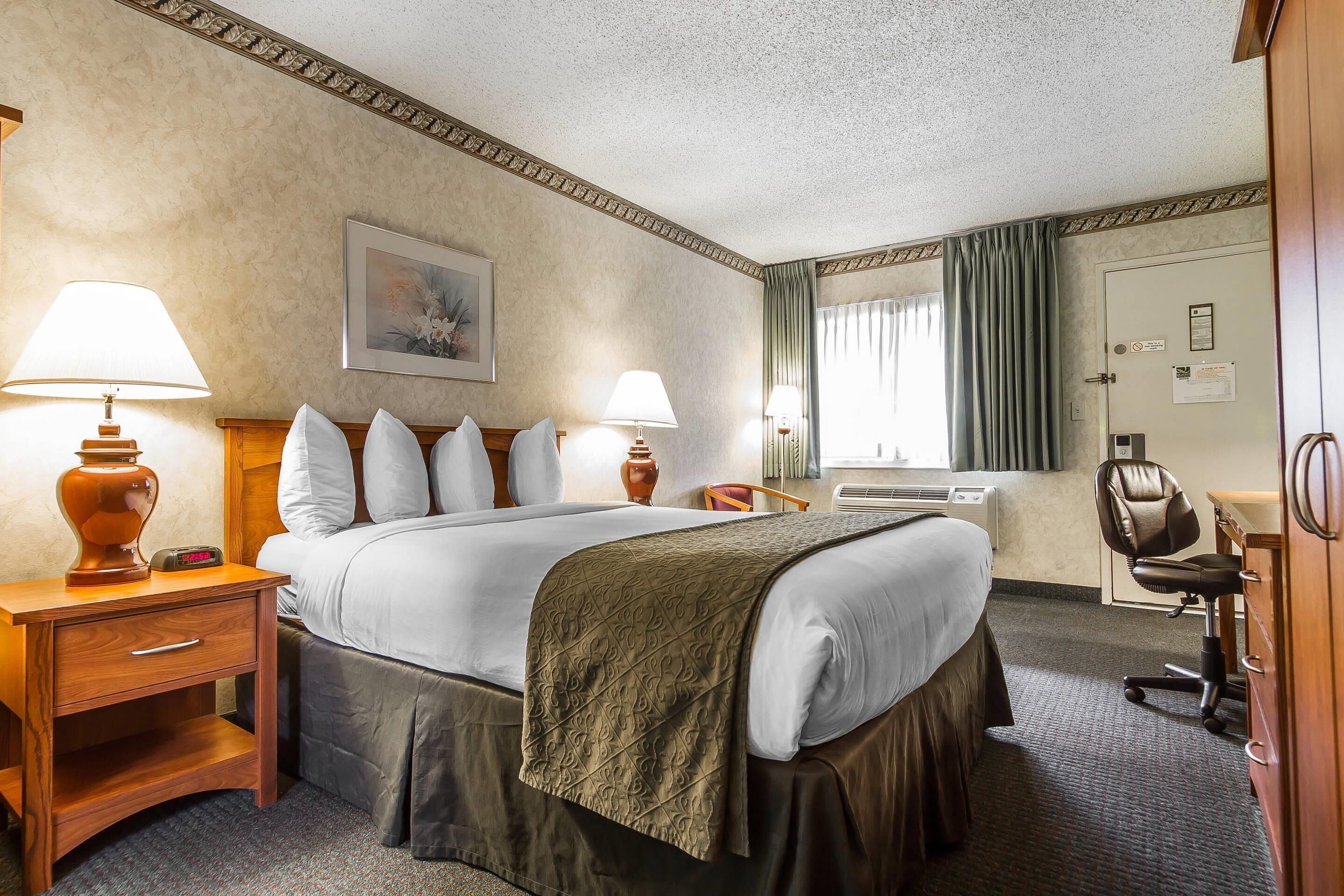 A hotel room with a large bed, two bedside lamps, a desk with a chair, a dresser, and a window with curtains.