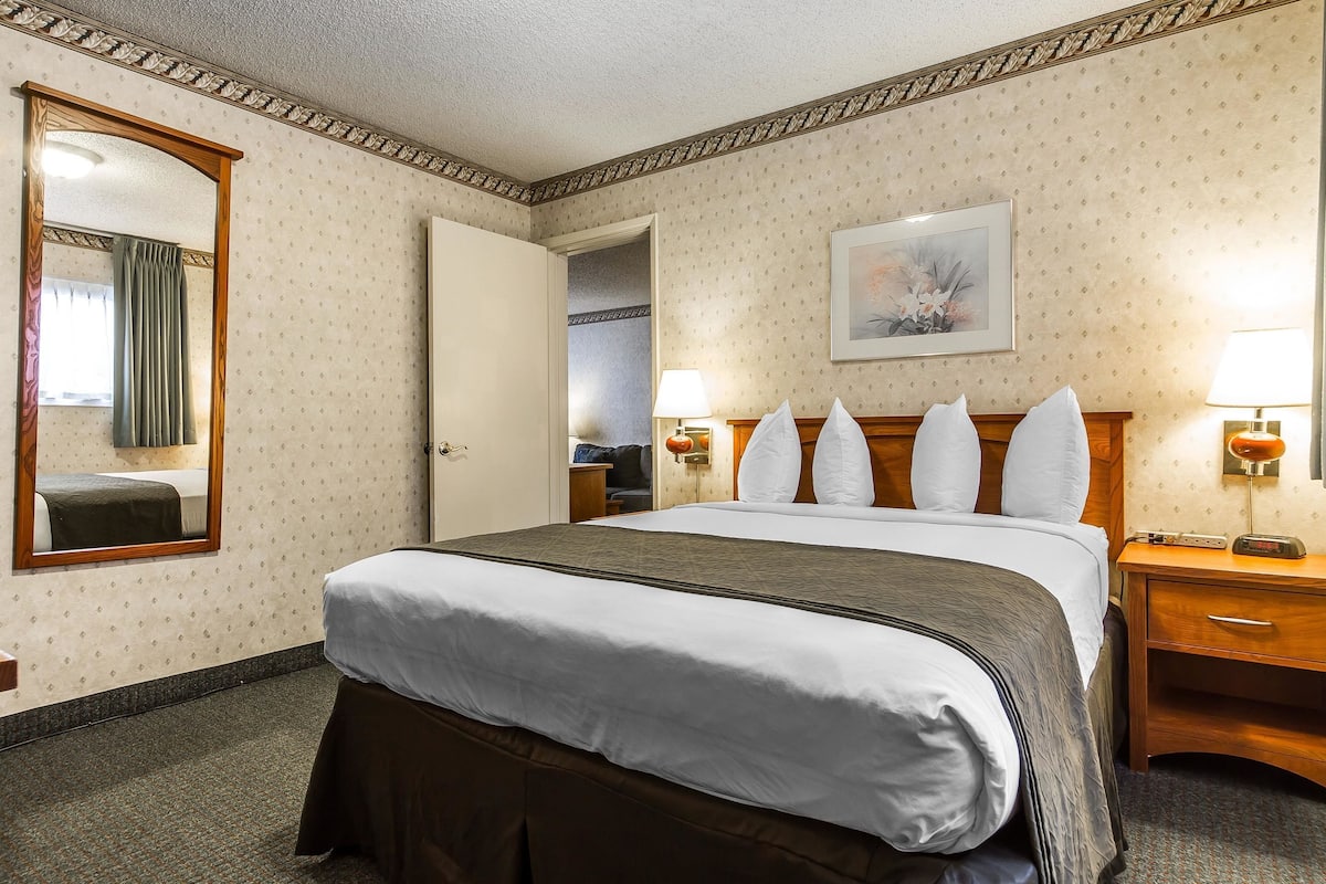Ein Hotelzimmer mit einem großen Bett, einem Holzkopfteil, einem Nachttisch mit Lampe, einem Spiegel und einem gerahmten Bild an der Wand.