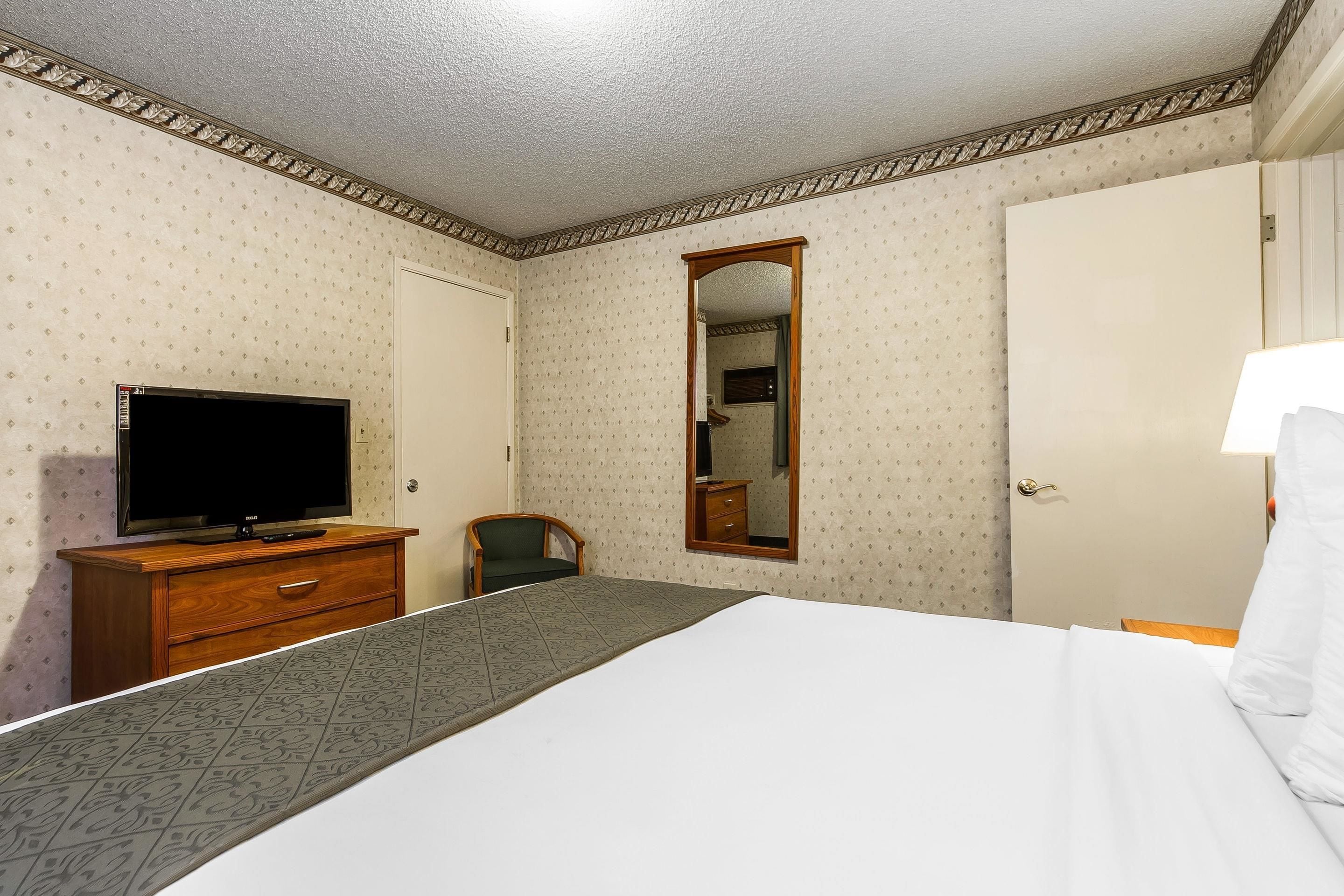 A hotel room with a bed, a television on a wooden dresser, a mirror, and a door.