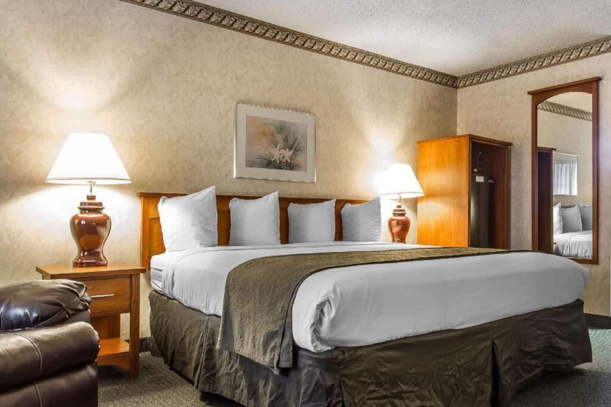 Ein Hotelzimmer mit einem Bett, zwei Nachttischlampen, einem Holznachttisch, einem Ledersessel, einem Bild an der Wand und einem Spiegel.