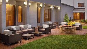 An outdoor patio area with a stone fire pit, cushioned seating, and a well-lit entrance with large windows and wall-mounted lights.