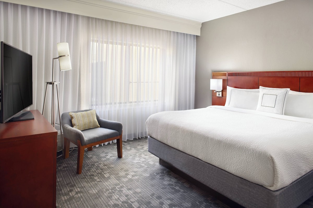 A hotel room with a large bed, a desk with a television, a chair, and a floor lamp.