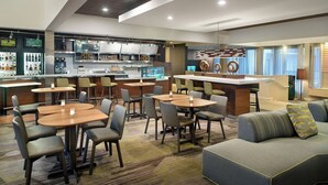 A modern hotel lobby with multiple seating areas, a bar, and a kitchen in the background.