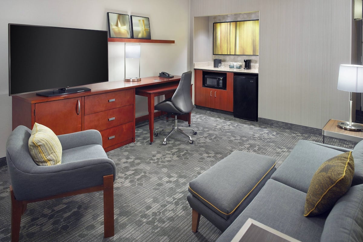 A hotel room with a flat-screen TV, a desk with a chair, a kitchenette, a sofa, and a lamp.