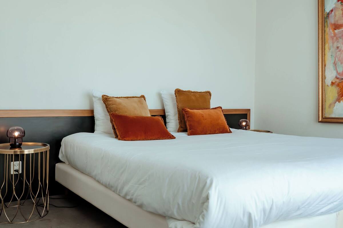 Ein ordentlich bezogenes Bett mit weißen Leinentüchern und zwei braunen Kissen, ein kleiner runder Tisch mit einer Lampe sowie ein Gemälde an der Wand.