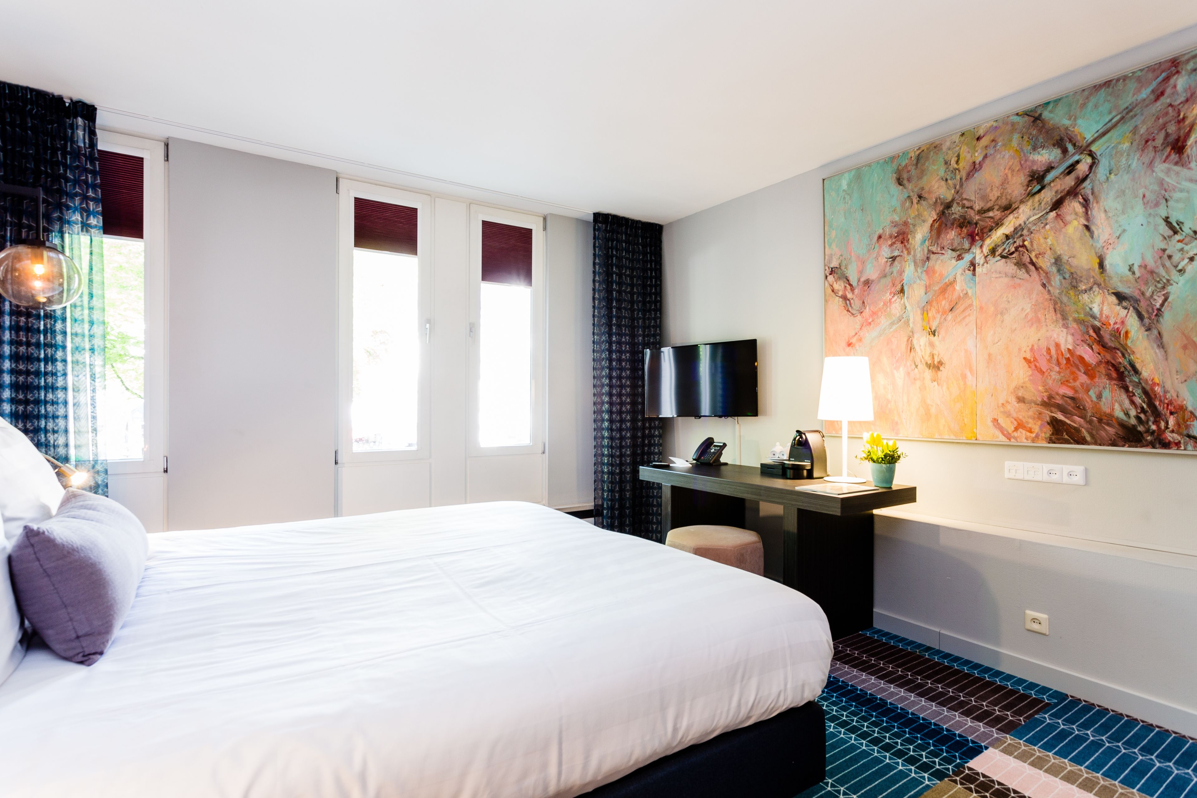 A hotel room with a large bed, a desk with a computer, a TV, and a colorful abstract painting on the wall.