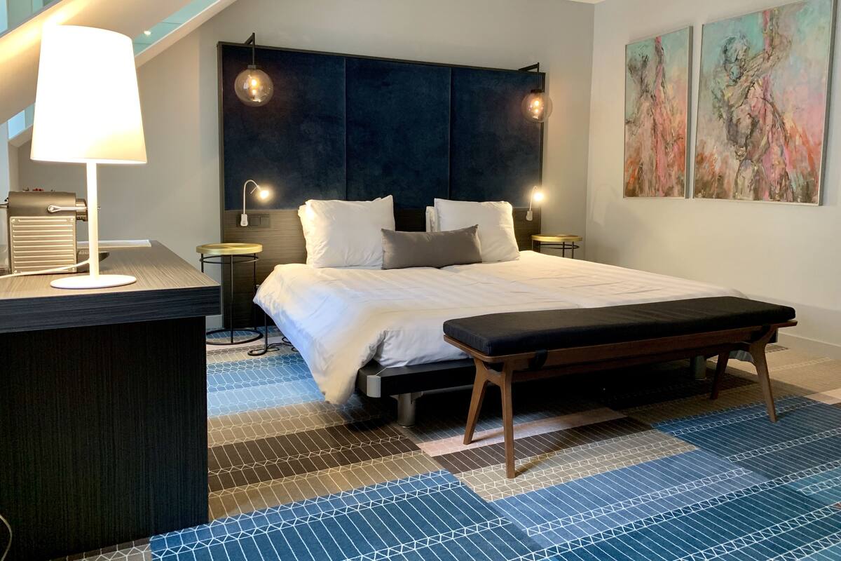 Ein modernes Schlafzimmer mit einem großen Bett, Nachttischlampen, einem Schreibtisch mit Lampe und abstrakter Wandkunst.
