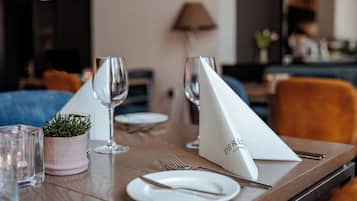 Mesa para comer servida con platos, cubiertos y vasos, con una planta en maceta y una servilleta blanca con texto.