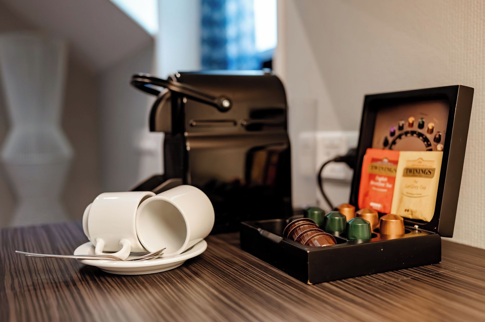 Una cafetera, dos tazas blancas, una bandeja con bolsitas de té y una caja de bombones sobre una mesa de madera.