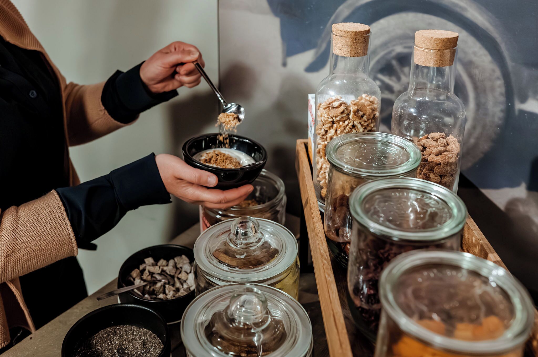 A person serving cereal from a bowl into a cup, with various food items in glass jars on a wooden tray.