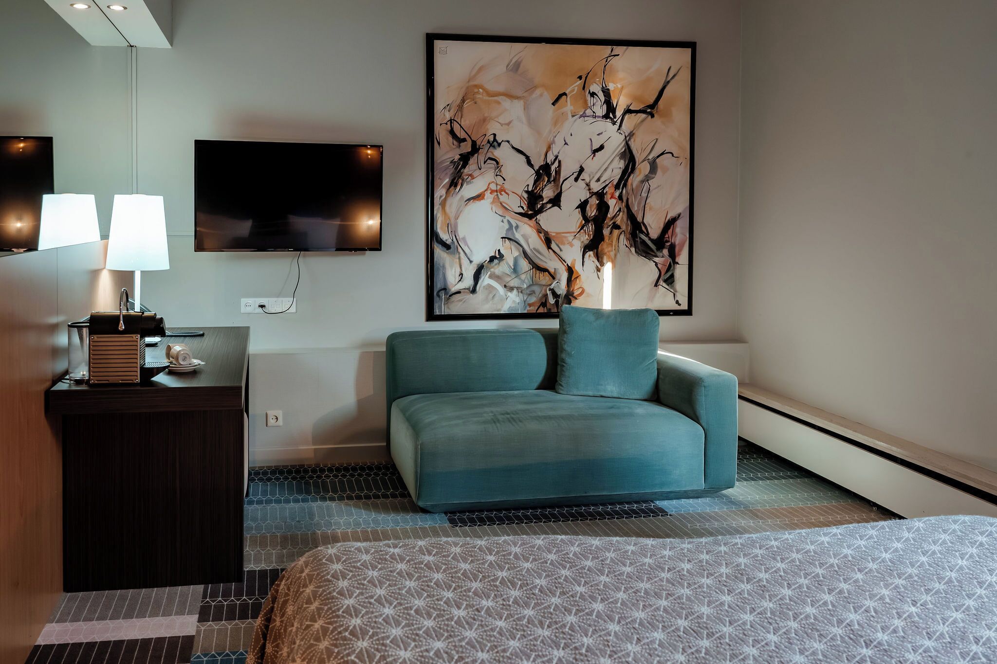 Hotelkamer met een bed, een bureau met een lamp, een televisie en een groot abstract schilderij.