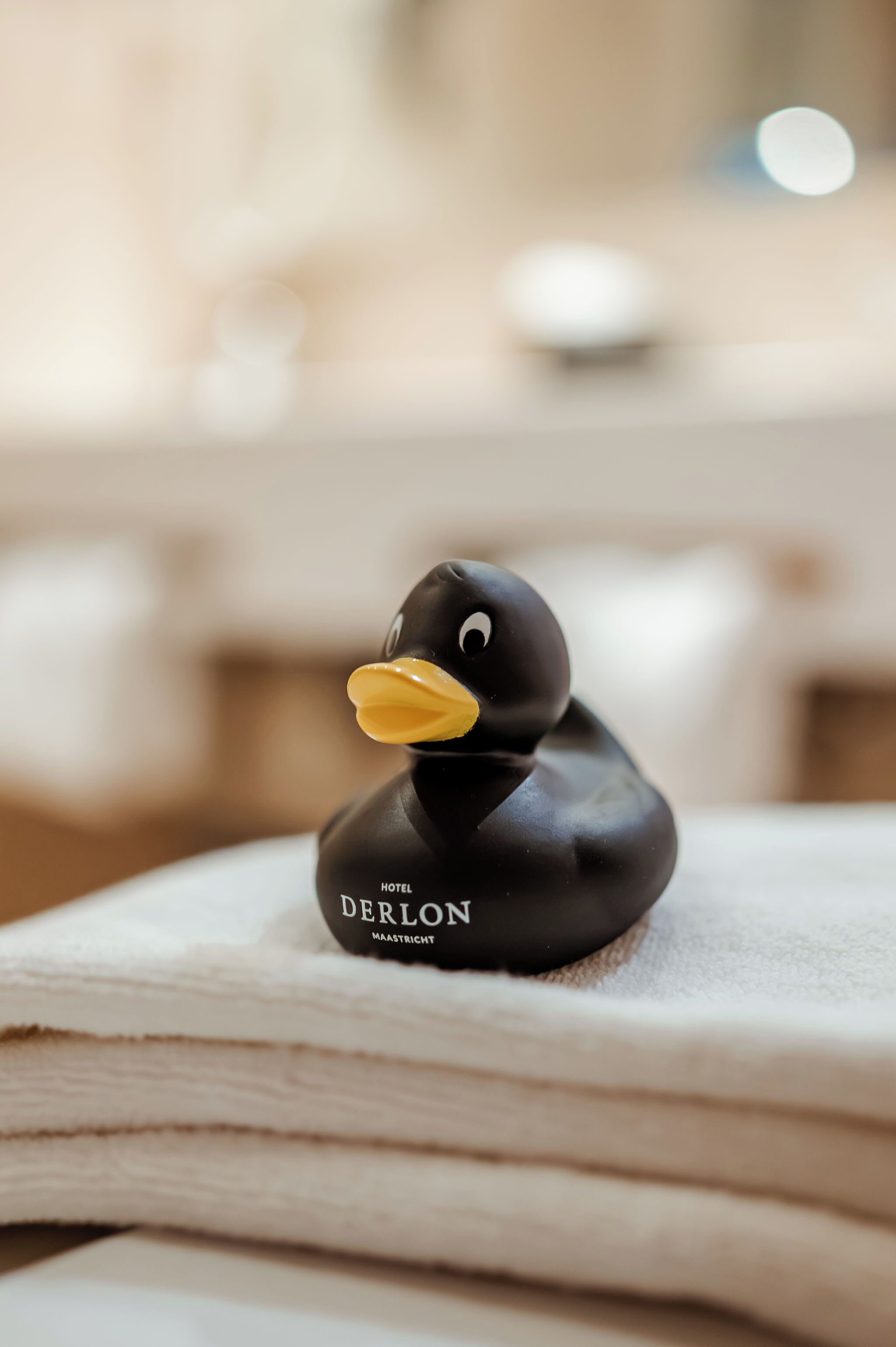 A black rubber duck with 'DERLON' and 'MAASRIET' text on it, placed on a stack of white towels.