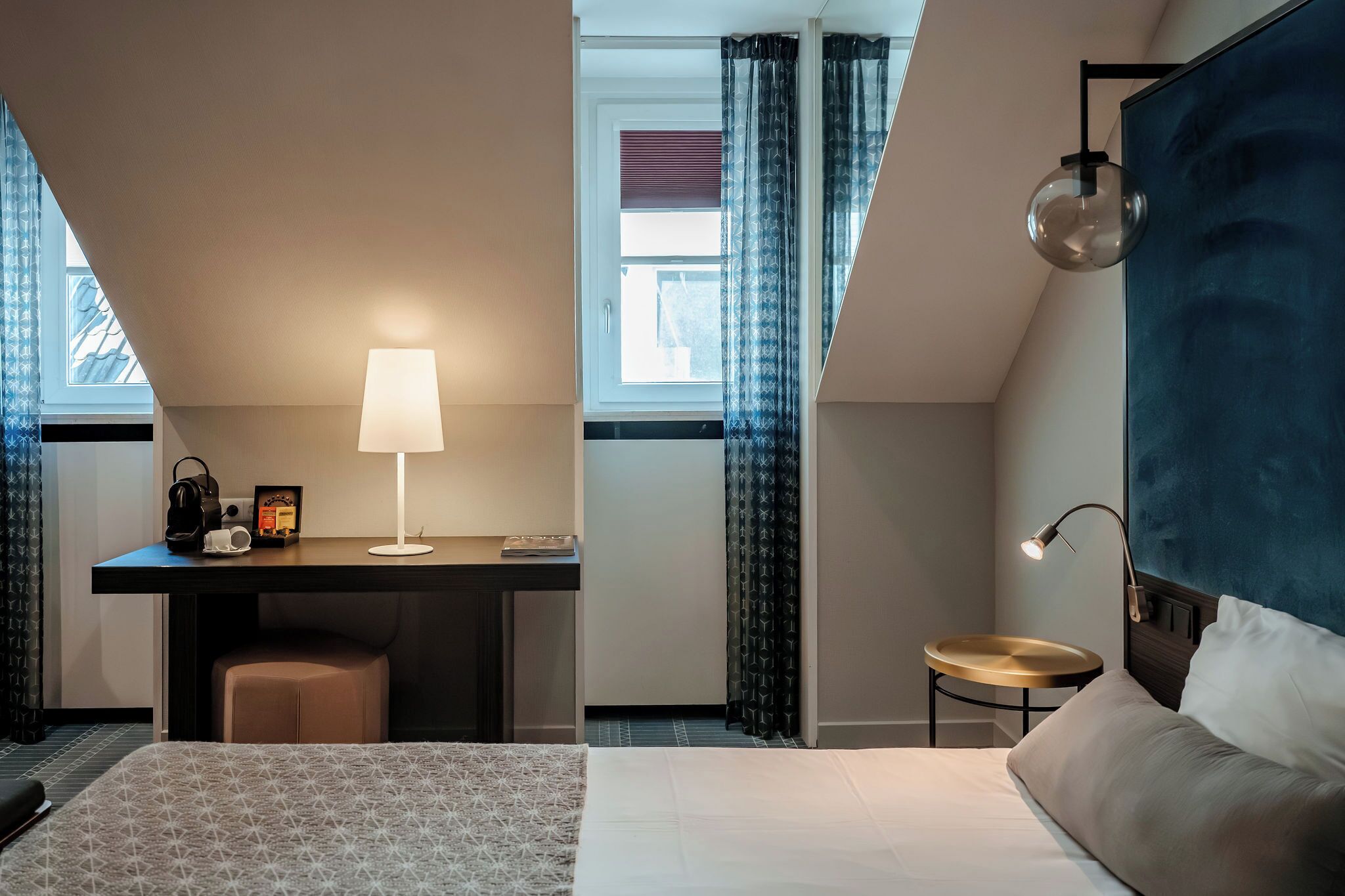 A hotel room with a bed, a desk with a lamp, a small round table, and a headboard.