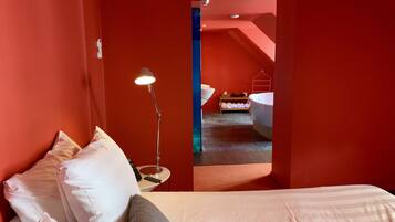 A hotel room with a bed, a desk lamp, and a view of a bathroom with a bathtub and towels.