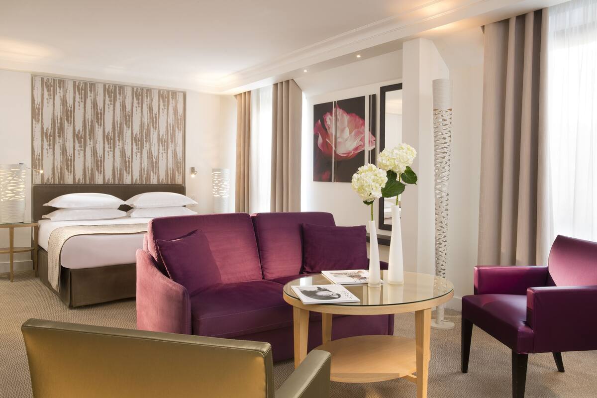 A hotel room with a bed, a purple sofa, a glass coffee table, and a chair. There is a vase with flowers on the table.