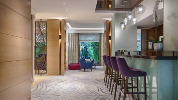 A modern hotel lobby with a patterned carpet, a bar area with purple stools, and a view of greenery through a large window.