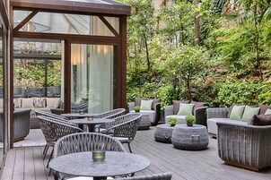 An outdoor patio with wicker furniture, a glass enclosure, and lush greenery.