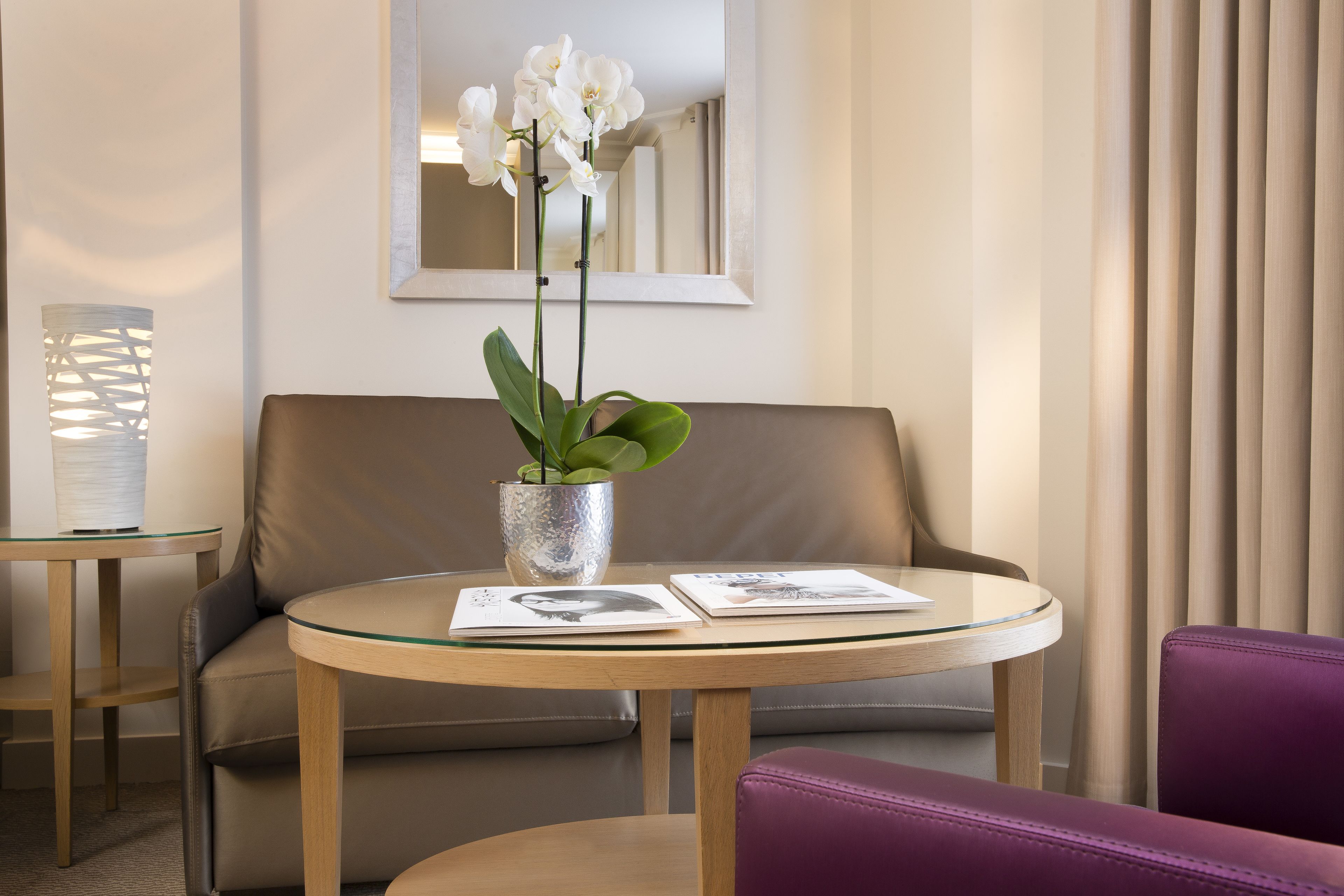 A modern hotel room with a round glass table, a leather sofa, a purple chair, and a potted orchid.