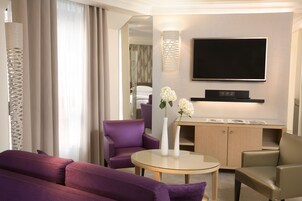 A modern hotel room with a purple sofa, a round glass table, a TV mounted on the wall, and a wooden cabinet.
