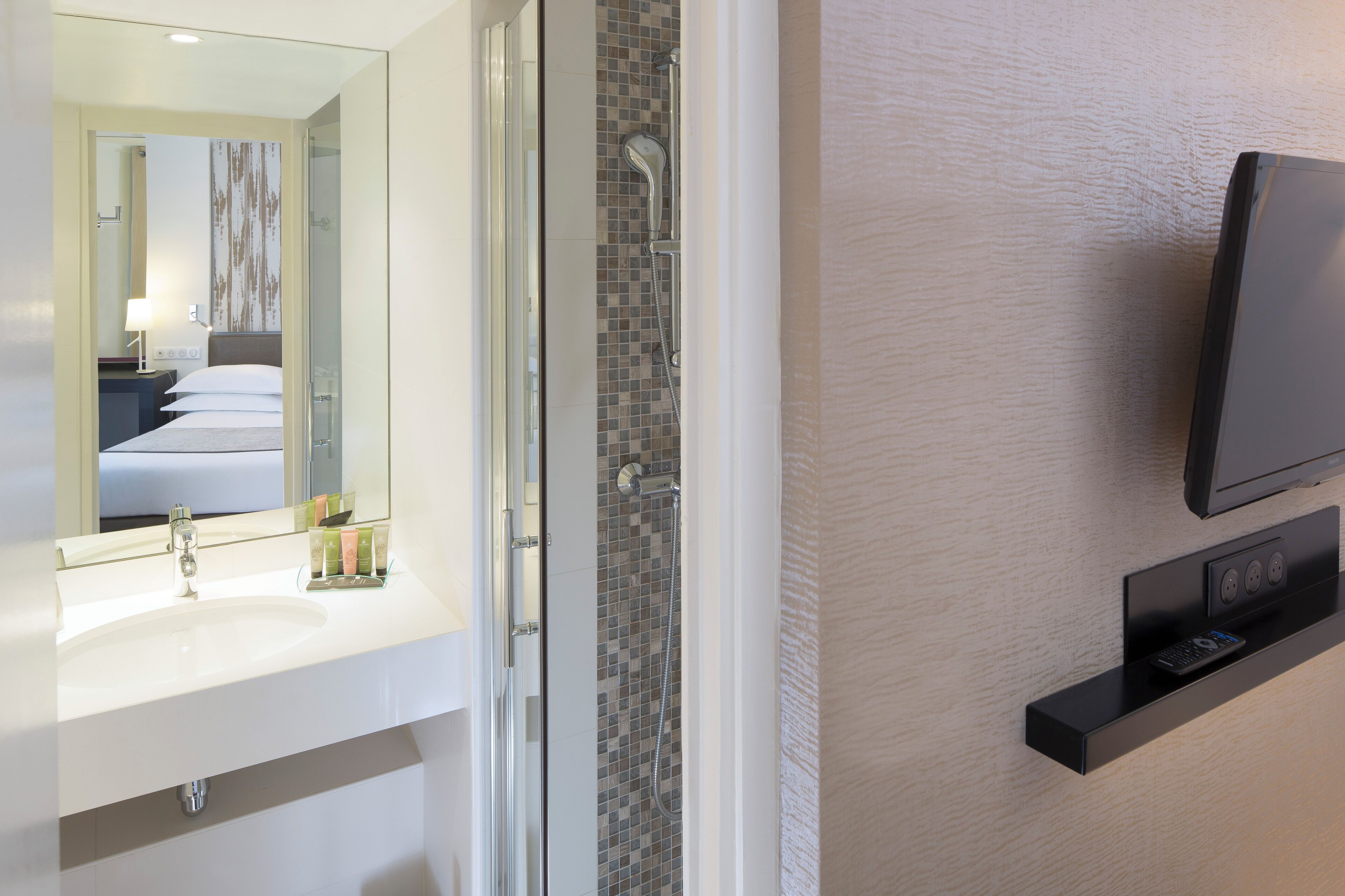 A modern bathroom with a glass shower enclosure, a white sink with a mirror, and a mounted television on the wall.