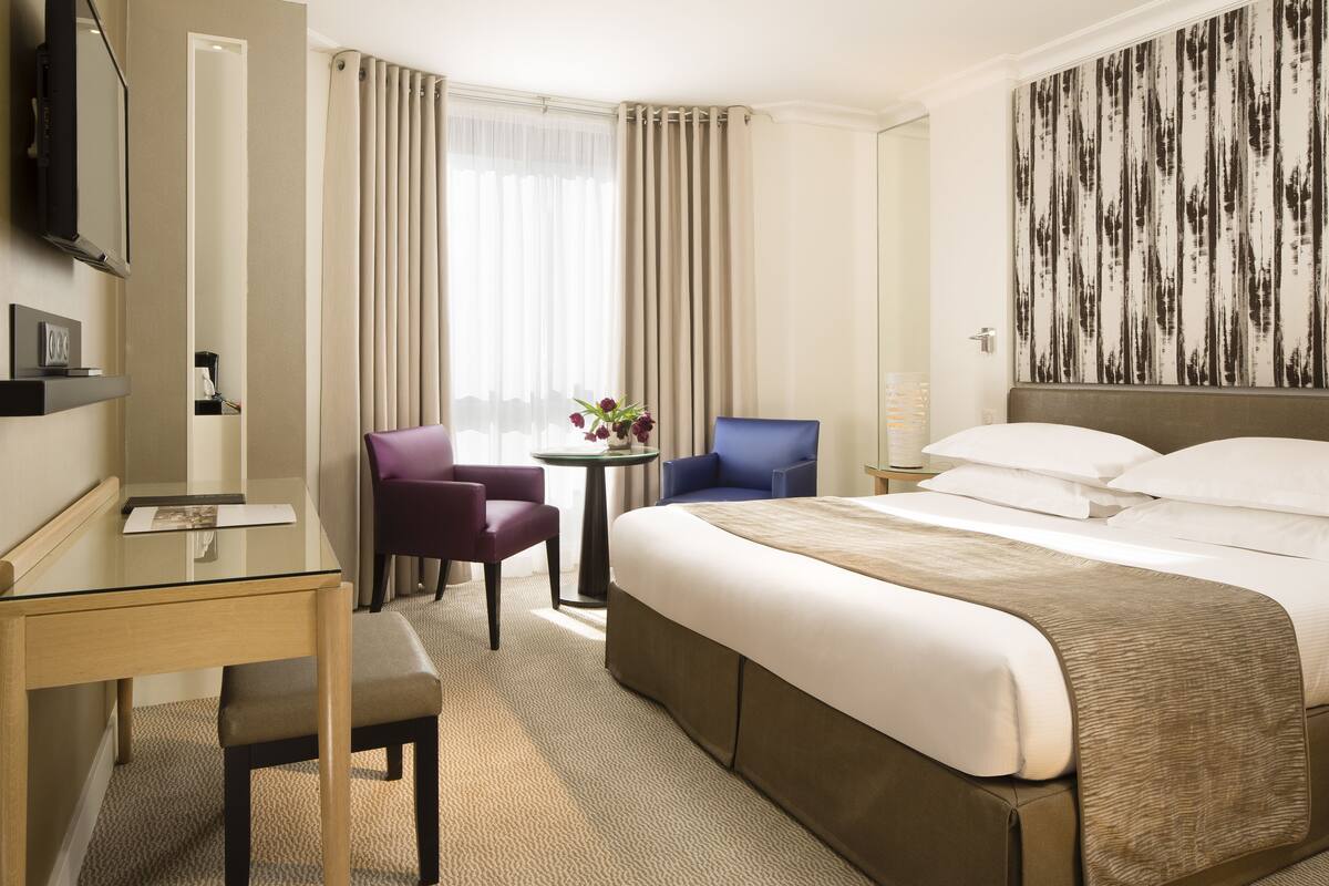 A hotel room with a large bed, a desk with a chair, a small table with a chair, and a large mirror.