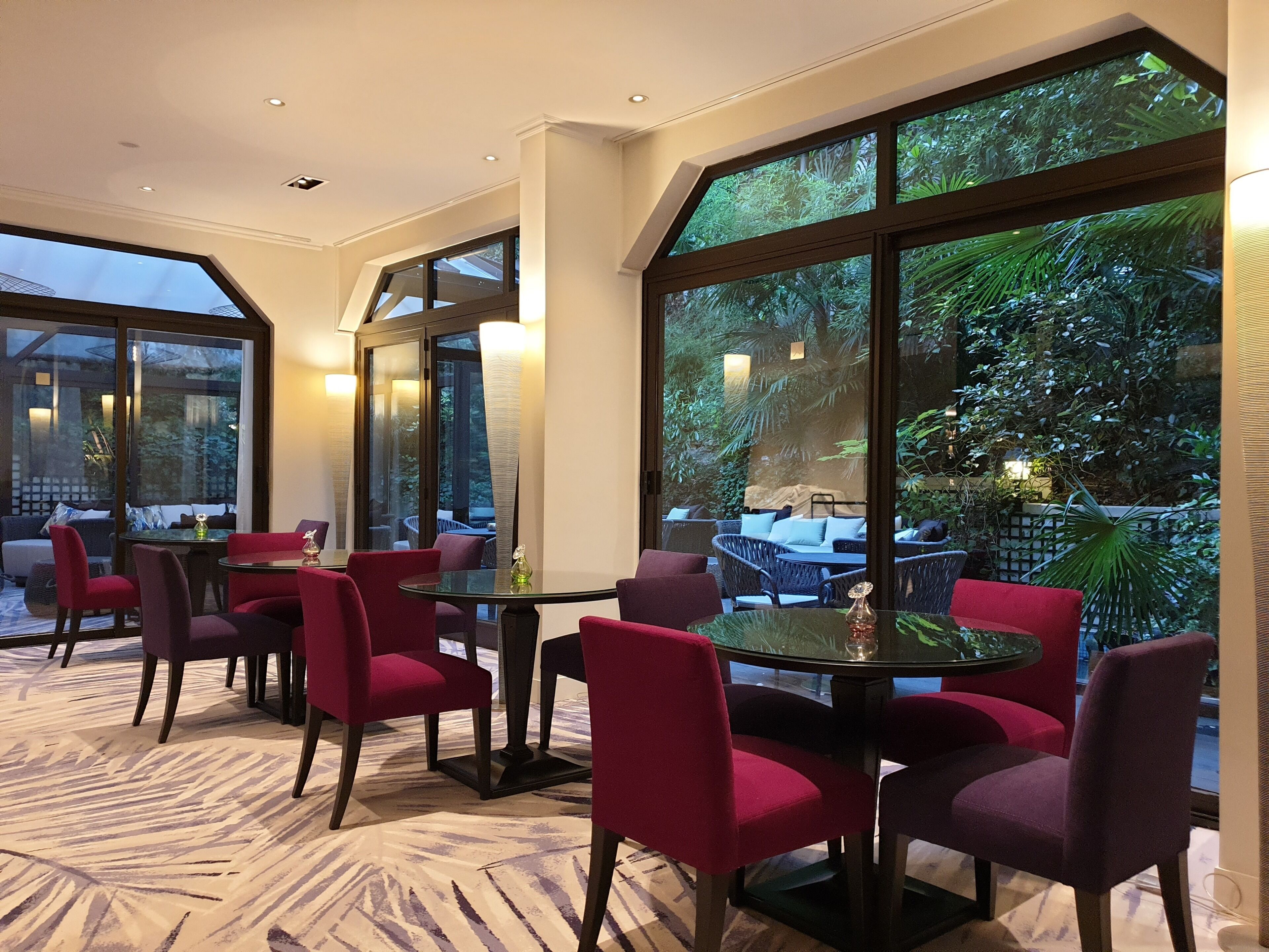 Um restaurante com mesas redondas e cadeiras vermelhas, janelas amplas com vista para o verde e um tapete estampado.