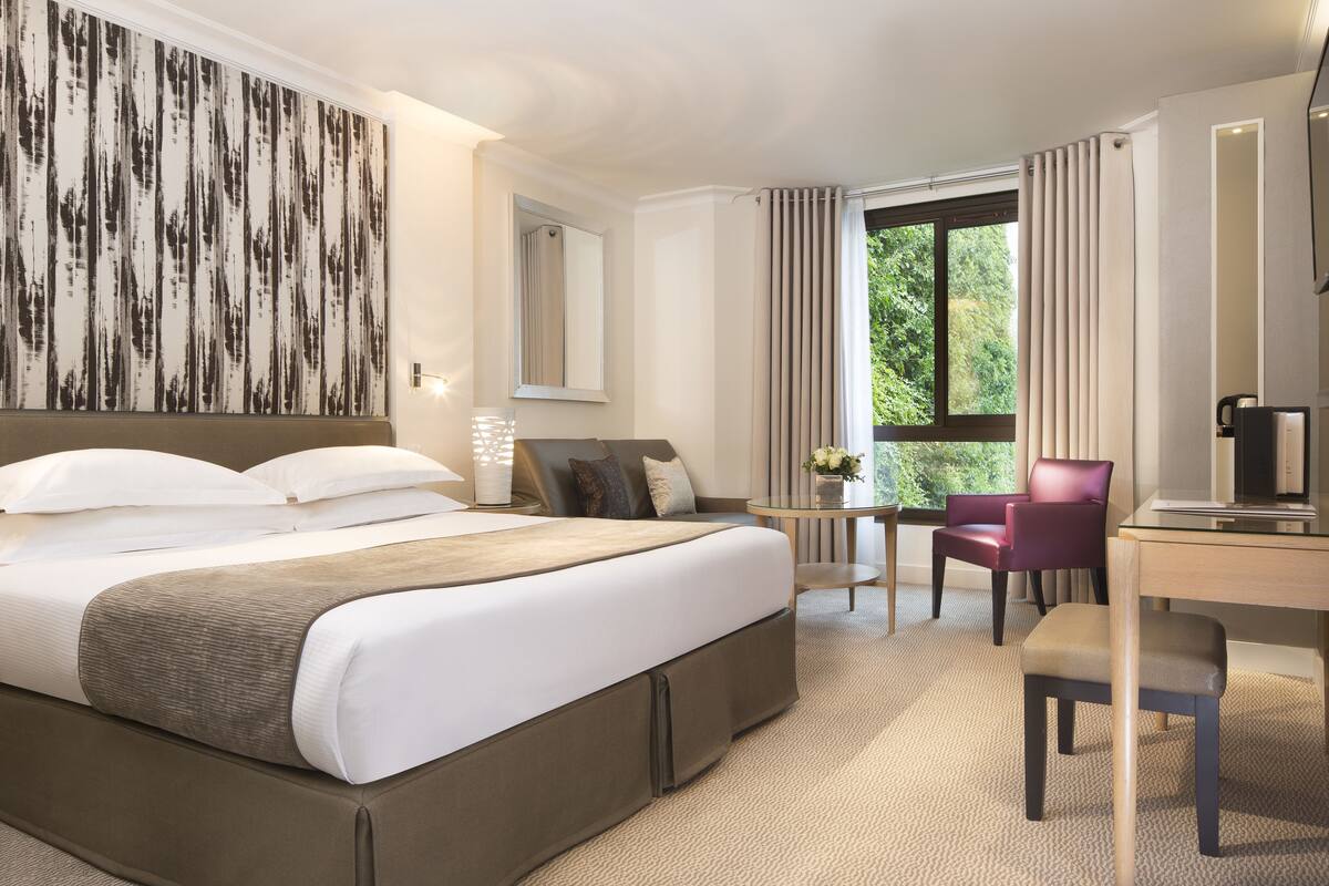 A modern hotel room with a large bed, a desk, a chair, and a window with curtains.