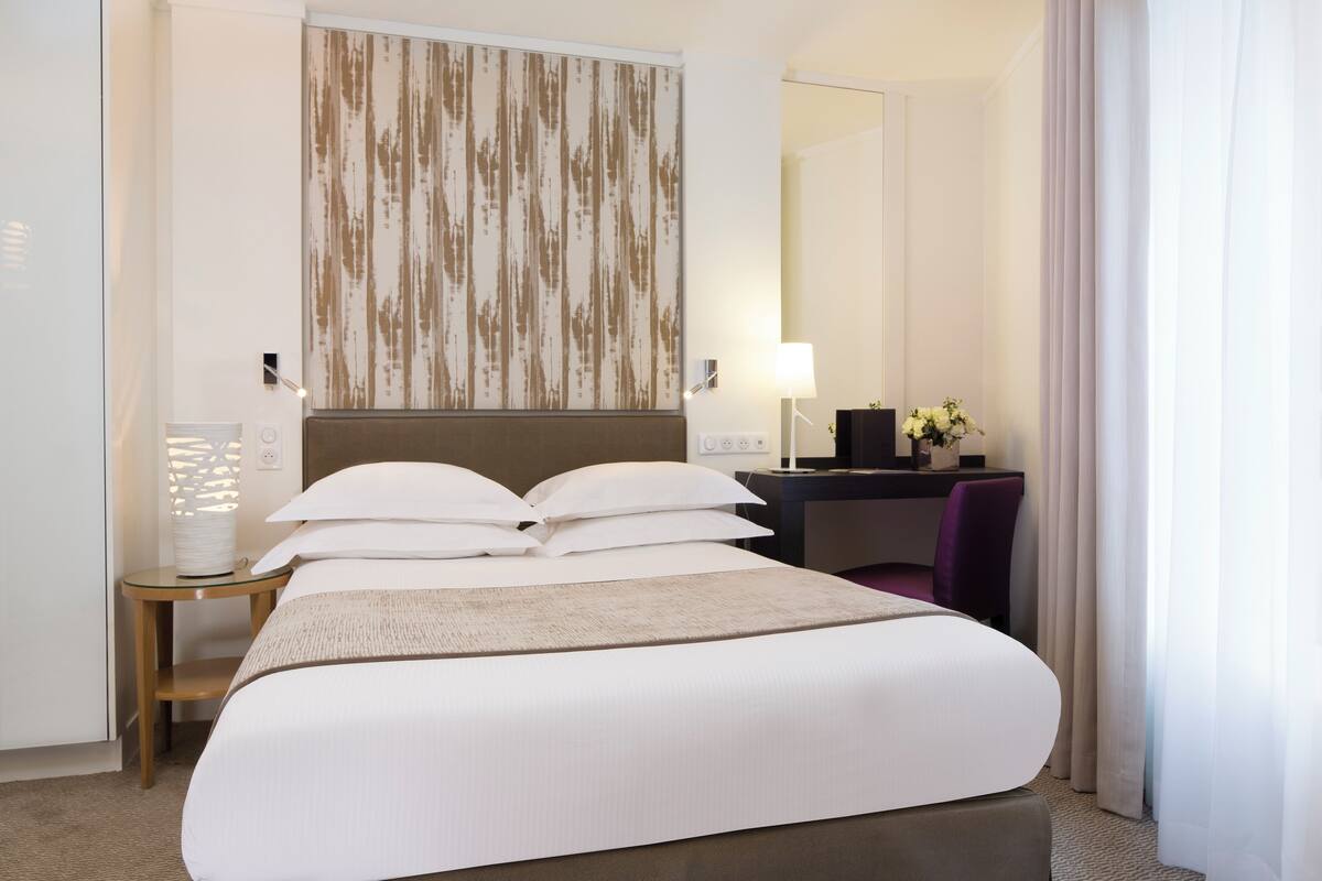 A hotel room with a large bed, a desk with a chair, a lamp, and a mirror.