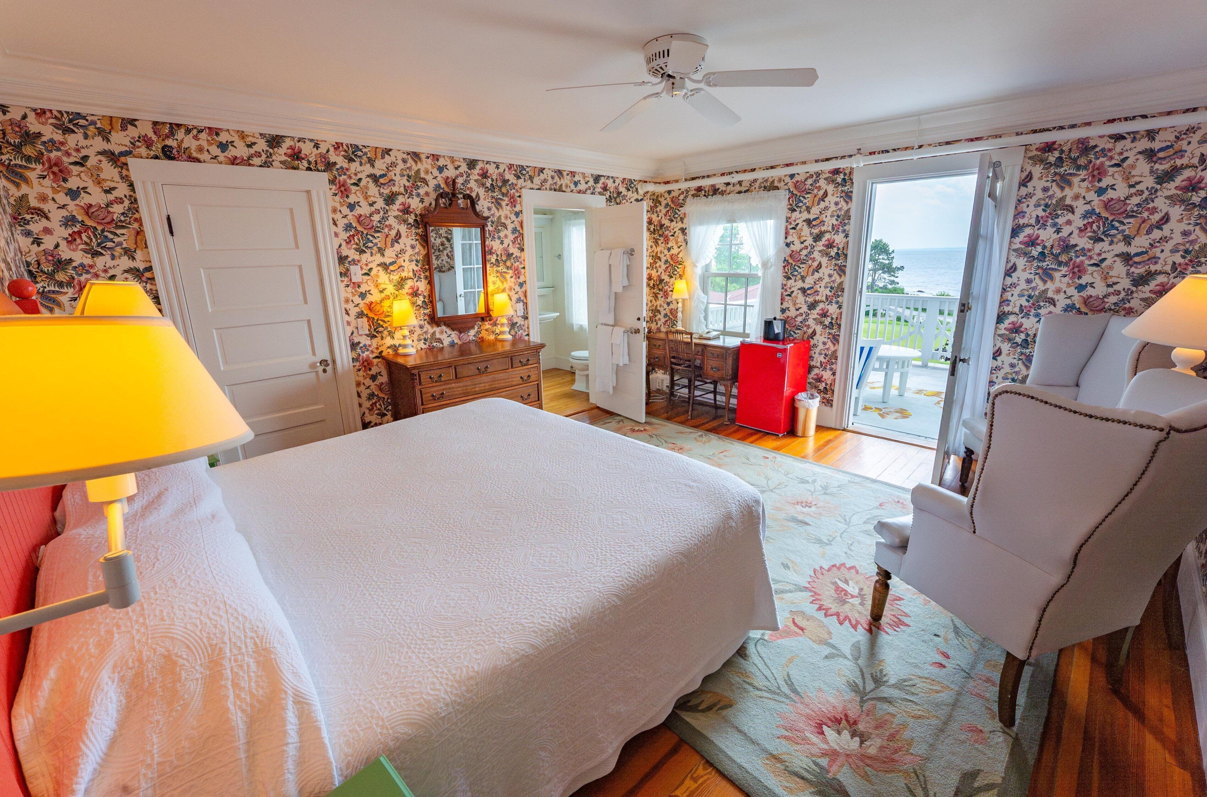 Une chambre à coucher comprenant un lit, un bureau, une chaise, une commode et une vue sur la mer, visible par la porte.