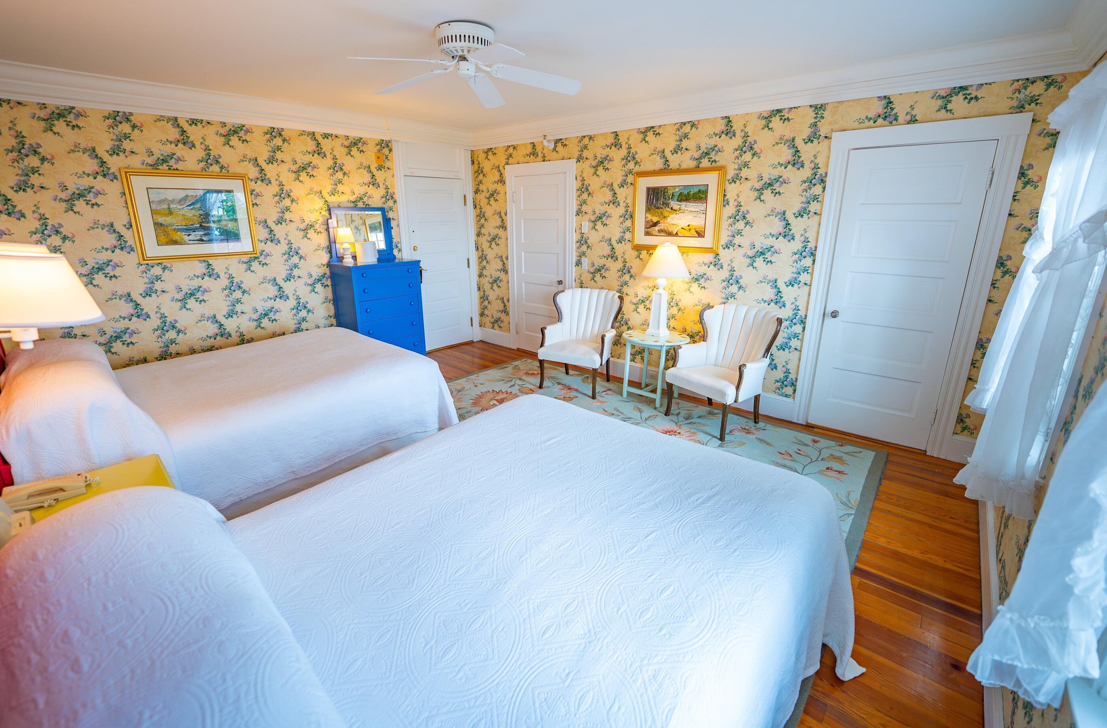 A hotel room with two beds, a blue dresser, a chair, a table with a lamp, and floral wallpaper.