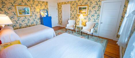 A hotel room with two beds, a blue dresser, a chair, a table with a lamp, and floral wallpaper.