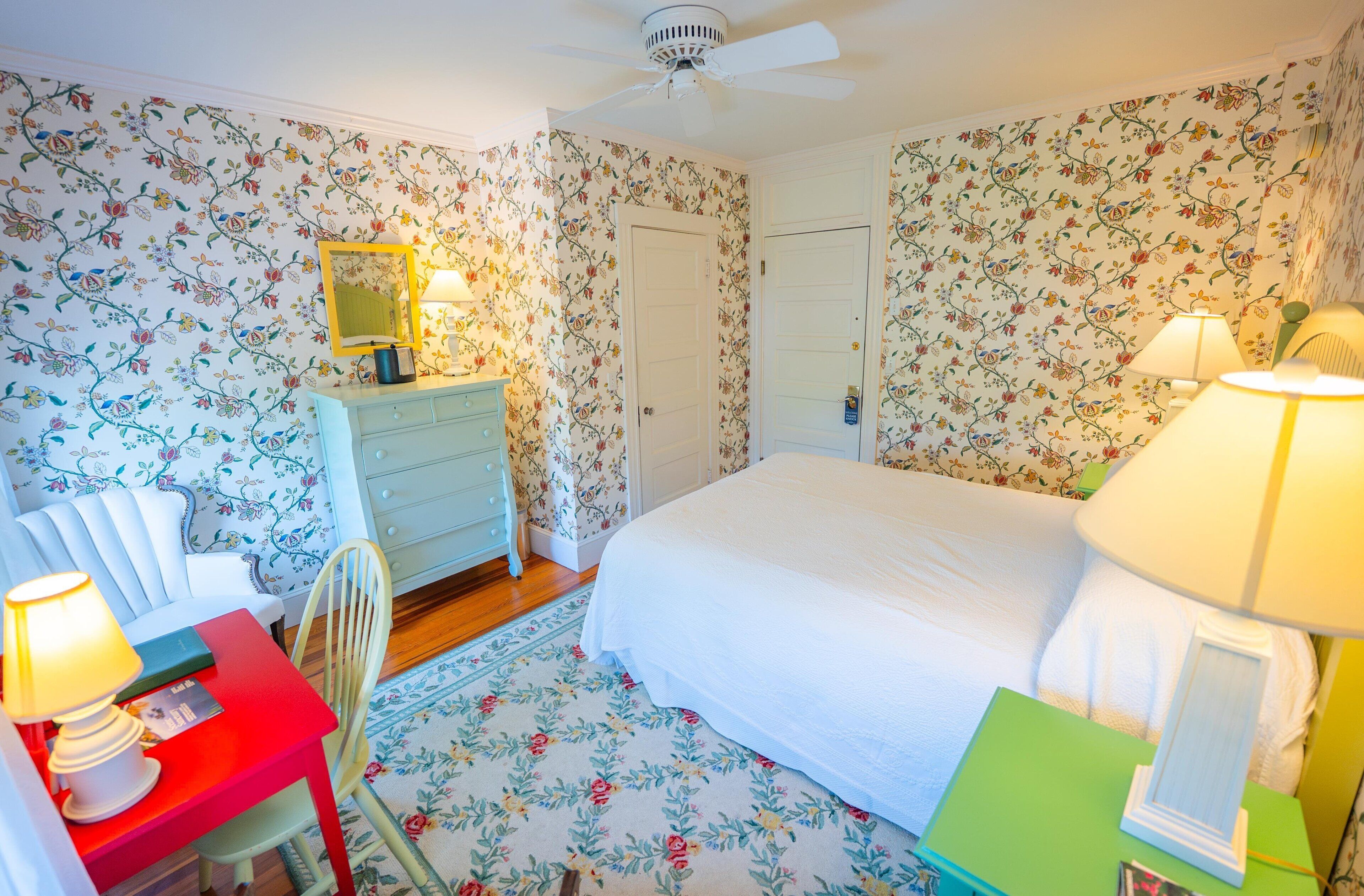 A bedroom with floral wallpaper, a bed, a dresser with a mirror, a desk with a chair, and two lamps.