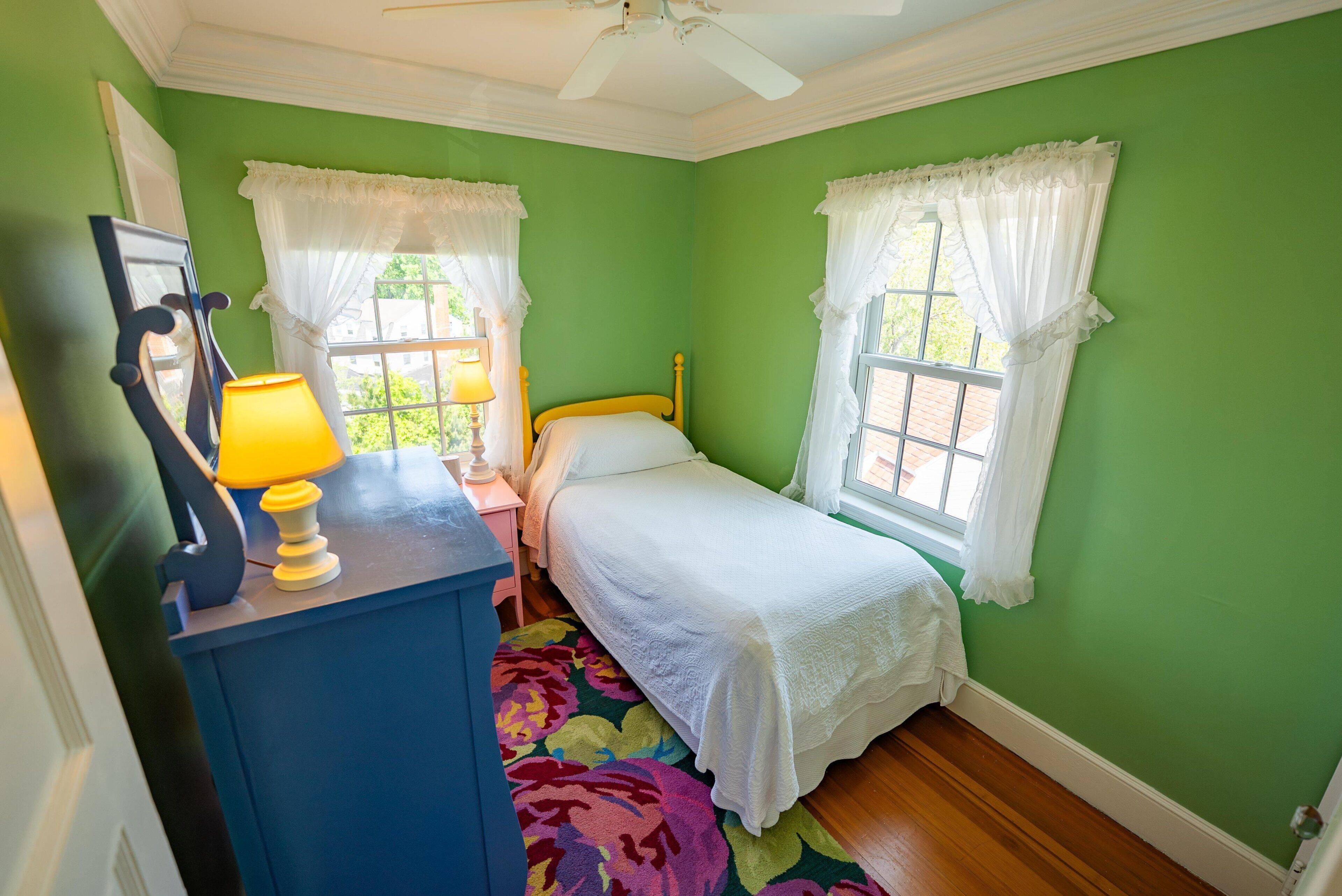 A bedroom with a bed, a dresser, a lamp, and two windows with white curtains.