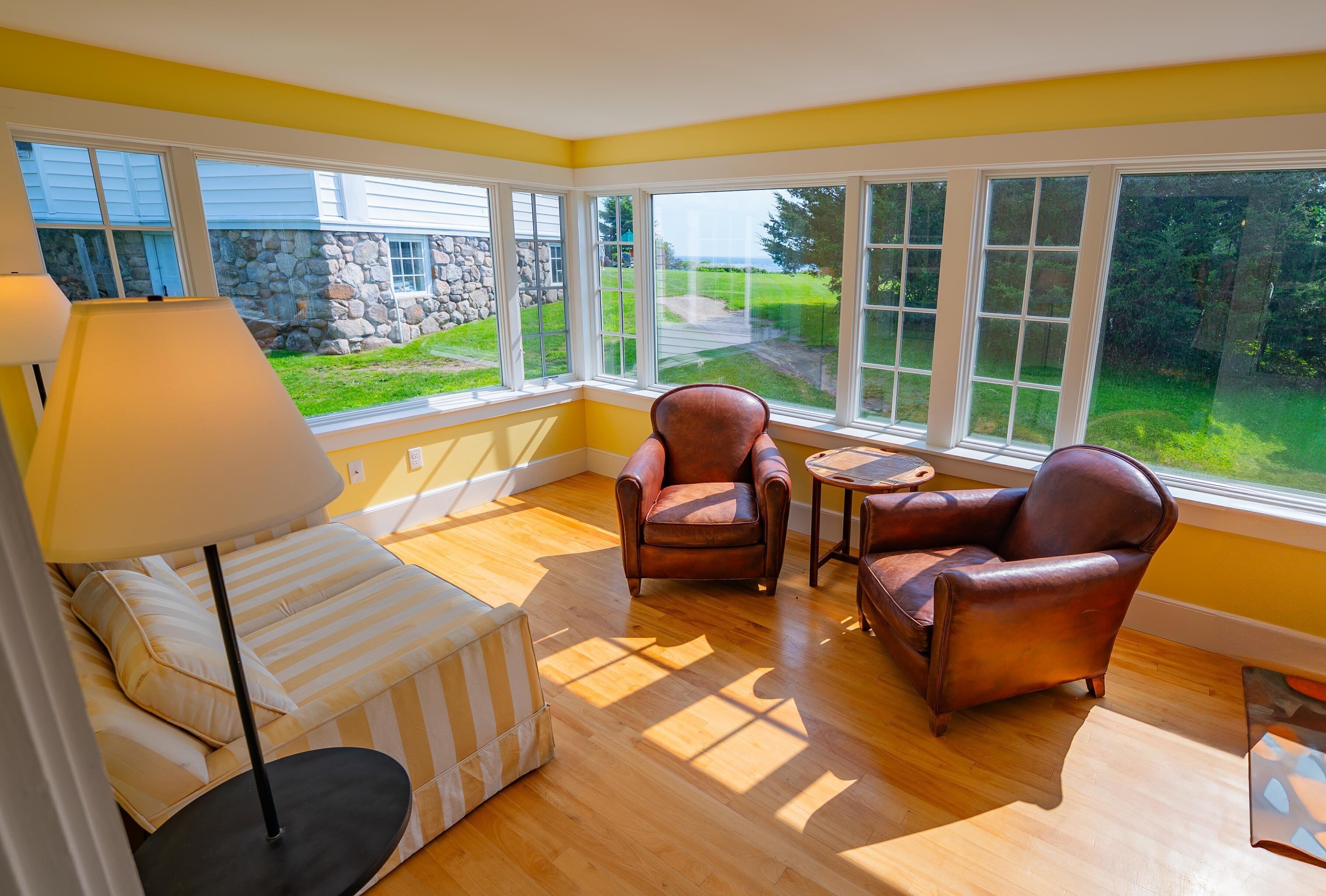A sunlit room with a sofa, two leather chairs, a small table, and a lamp. Large windows offer a view of a stone wall and greenery outside.